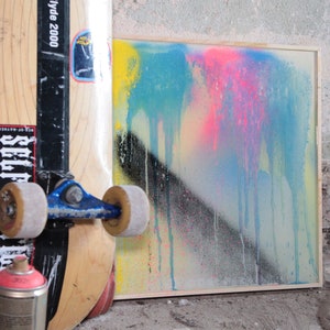 Könnte beinhalten: Ein gerahmtes abstraktes Gemälde mit Tropfen blauer, rosa, gelber und schwarzer Farbe auf weißem Hintergrund. Ein Skateboard mit Rädern und einem Aufkleber mit der Aufschrift "Hyde 2000" lehnt an dem Gemälde.