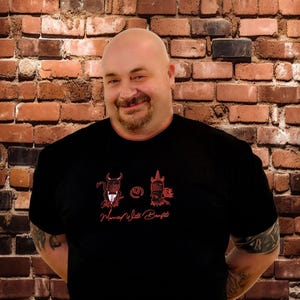 May include: A black t-shirt with a red graphic design and the text "Married With Benefits". The design features two cartoon figures. The background is a brick wall.