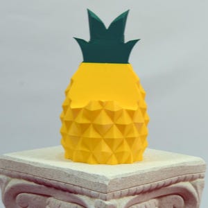 May include: A yellow pineapple sculpture with a green top sits atop a decorative stone pedestal. The pineapple has a geometric design and is brightly colored. The pedestal is a light beige color with carved details. The background is a plain white.