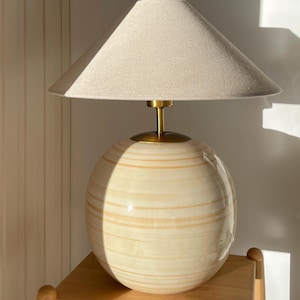 Puede incluir: Una l&aacute;mpara de mesa de cer&aacute;mica beige con una base redonda y una pantalla de lino. La l&aacute;mpara tiene un acento de metal dorado en la base y un remate de metal dorado en la parte superior de la pantalla.