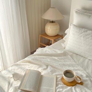 Puede incluir: Una cama blanca con una colcha de crochet blanca, un libro y una taza de caf&eacute; sobre un posavasos de madera. Una l&aacute;mpara con una pantalla beige y una base beige se encuentra sobre una mesita de noche de madera.
