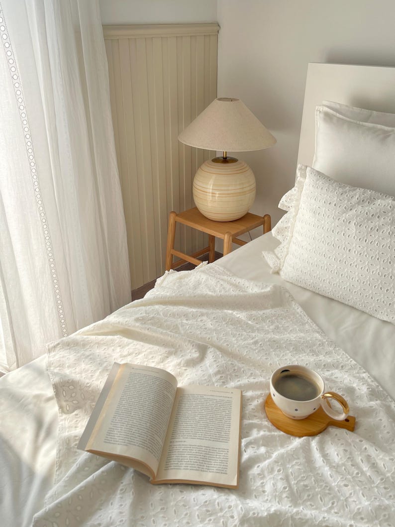 Puede incluir: Una cama blanca con una colcha de crochet blanca, un libro y una taza de caf&eacute; sobre un posavasos de madera. Una l&aacute;mpara con una pantalla beige y una base beige se encuentra sobre una mesita de noche de madera.