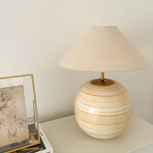 Puede incluir: Una l&aacute;mpara de mesa blanca y dorada con una pantalla de lino beige. La base de la l&aacute;mpara es una esfera redonda con rayas doradas. La l&aacute;mpara est&aacute; sobre una mesa blanca con tres libros apilados debajo.
