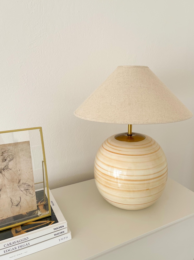 Puede incluir: Una l&aacute;mpara de mesa blanca y dorada con una pantalla de lino beige. La base de la l&aacute;mpara es una esfera redonda con rayas doradas. La l&aacute;mpara est&aacute; sobre una mesa blanca con tres libros apilados debajo.