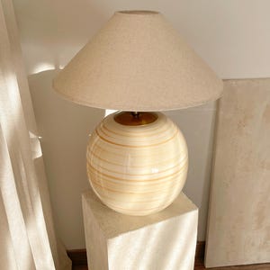 Puede incluir: Una l&aacute;mpara de mesa de cer&aacute;mica beige con una base redonda y una pantalla de lino blanca. La l&aacute;mpara est&aacute; sobre un pedestal blanco.