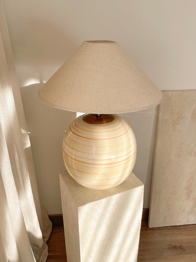 Puede incluir: Una l&aacute;mpara de mesa de cer&aacute;mica beige con una base redonda y una pantalla de lino blanca. La l&aacute;mpara est&aacute; sobre un pedestal blanco.