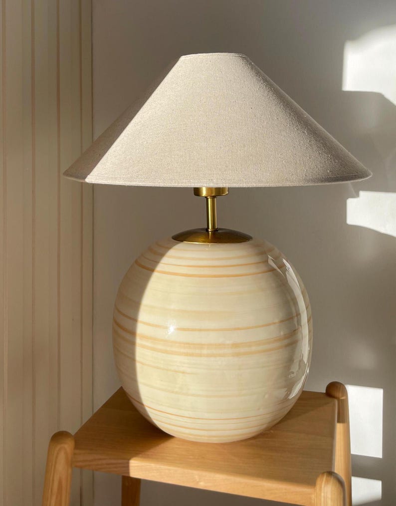 Puede incluir: Una l&aacute;mpara de mesa de cer&aacute;mica beige con una base redonda y una pantalla de lino. La l&aacute;mpara tiene un acento de metal dorado en la base y un remate de metal dorado en la parte superior de la pantalla.