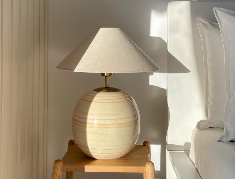 Puede incluir: Una l&aacute;mpara de mesa de cer&aacute;mica beige con una base redonda y una pantalla de lino blanca. La l&aacute;mpara est&aacute; sobre una mesa auxiliar de madera.