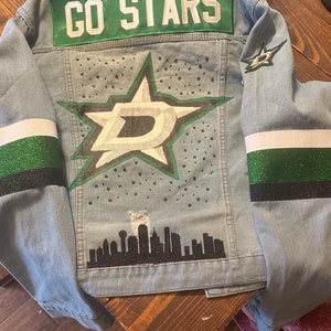 May include: A blue denim jacket with green and white stripes on the sleeves. The back of the jacket features a large green star with a white "D" inside, surrounded by small green dots. The text "GO STARS" is on the top of the jacket. A black silhouette of a city skyline is at the bottom of the jacket.