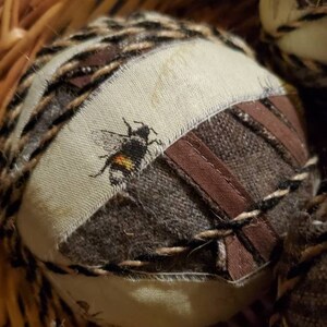 May include: A brown wicker basket filled with round, fabric-covered ornaments. Each ornament features a different pattern and a small embroidered bee.