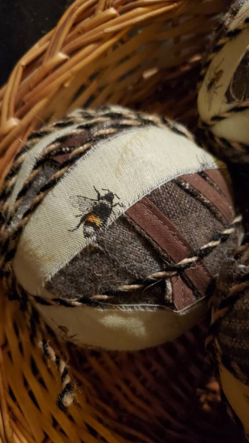 May include: A brown wicker basket filled with round, fabric-covered ornaments. Each ornament features a different pattern and a small embroidered bee.