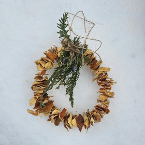May include: A handmade wreath composed of dried orange slices, evergreen sprigs, and twine, set against a snowy backdrop. The wreath is circular, with the orange slices forming the main body. A natural, rustic decoration.