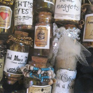 May include: A collection of small glass bottles with various labels and contents. The labels include "EWT'S EYES", "Dead Man's T", "Spooks Delight", "maggots", and "Ghost in a bowl".