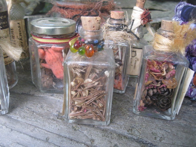 May include: Six small glass jars with cork tops filled with various dried botanicals and other items. The jars are arranged in a row on a wooden surface. The jars are decorated with twine and other embellishments.