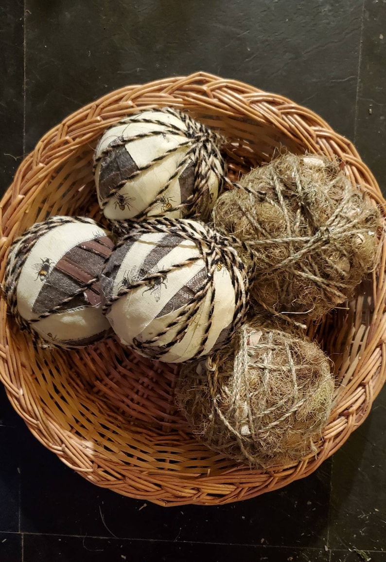 May include: Four round ornaments in a wicker basket. Two are covered in fabric with a bee pattern and wrapped with twine. The other two are wrapped in twine and covered in brown fibers.