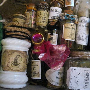 May include: A collection of small glass bottles with various labels and contents. Some bottles are tied with twine and have cork stoppers. The labels include text such as "EWT'S EYES", "Spook's Brew", and "Poison Ivy".