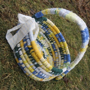 Può includere: Un cesto intrecciato con ritagli di tessuto in tonalità di blu, giallo e bianco. Il cesto ha un manico e un fiocco fatto di tessuto bianco con strisce blu.