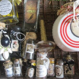 May include: A collection of small glass bottles with various labels and contents, including "Vampire Fangs", "Crow Nails", "Ink Tail", "Dried Maggot", and "Snake Root". The bottles are arranged in a wicker basket with a white and red striped lid.