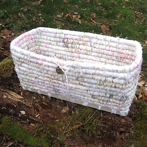 May include: Rectangular woven basket with a white and floral fabric design. The basket has a light-colored wooden base and a small, round, bronze-colored charm attached with a white string. The basket is sitting on a log.
