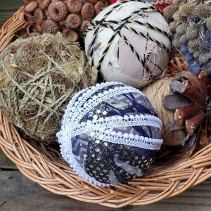 May include: A wicker basket filled with various decorative ornaments, including a brown acorn cluster, a ball wrapped in twine, a white ball with black twine, a red and blue patterned ball, a blue and white ball with sequins and lace, and a brown pine cone.