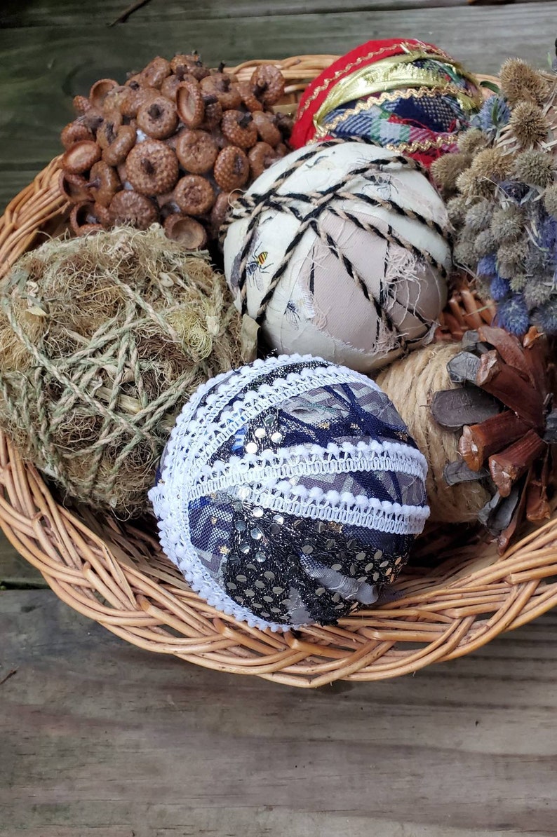 May include: A wicker basket filled with various decorative ornaments, including a brown acorn cluster, a ball wrapped in twine, a white ball with black twine, a red and blue patterned ball, a blue and white ball with sequins and lace, and a brown pine cone.