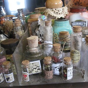 May include: A collection of small glass bottles with cork stoppers and labels. The bottles are filled with various liquids and have a vintage, apothecary aesthetic. Some of the labels include text such as "Graveyard Fog" and "Bewitching Potion".