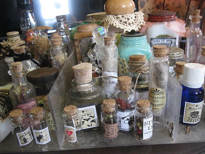 May include: A collection of small glass bottles with cork stoppers and labels. The bottles are filled with various liquids and have a vintage, apothecary aesthetic. Some of the labels include text such as "Graveyard Fog" and "Bewitching Potion".