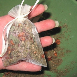 May include: A white sheer pouch filled with dried herbs and flowers. The pouch is held in a hand.