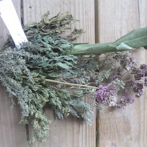 May include: A bundle of dried herbs and flowers, including thyme and oregano, with a large green leaf. The herbs are tied together with a natural twine. The background is a wooden surface.