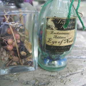 May include: Two glass bottles with labels. The bottle on the left contains dried herbs and berries. The bottle on the right has a label that reads "Enchantment Additive Eye of Newt" with a black raven illustration.