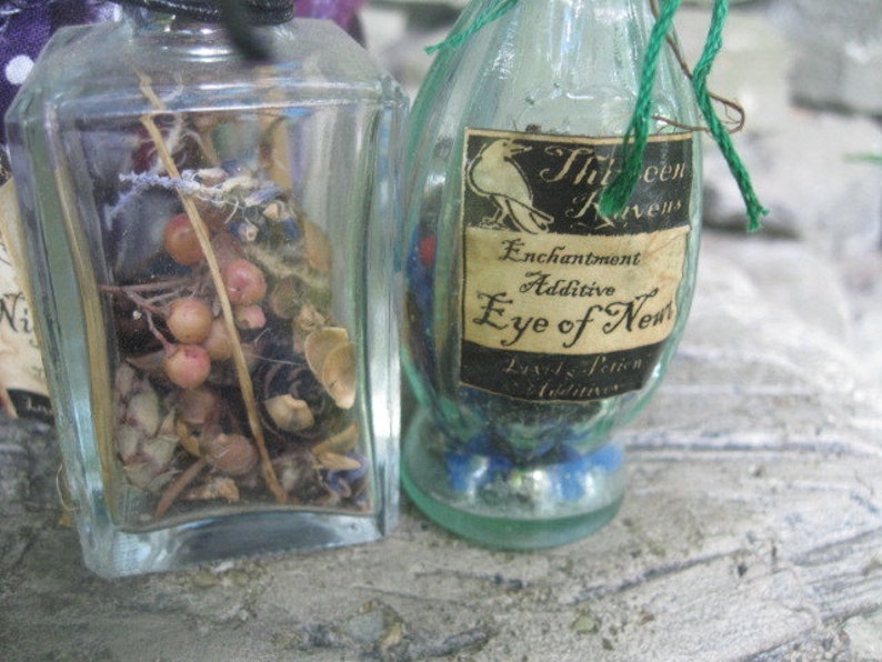 May include: Two glass bottles with labels. The bottle on the left contains dried herbs and berries. The bottle on the right has a label that reads "Enchantment Additive Eye of Newt" with a black raven illustration.