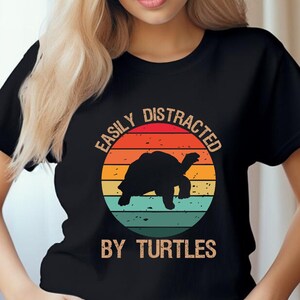 May include: Black t-shirt with a circular graphic featuring a turtle silhouette against a sunset design in orange, yellow, and teal. The text "EASILY DISTRACTED BY TURTLES" is printed around the circle.
