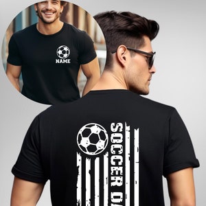 May include: Black t-shirt with a soccer ball graphic and the word "NAME" below it. The back of the shirt features a soccer ball and the words "SOCCER DAD" in a vertical design resembling a flag.