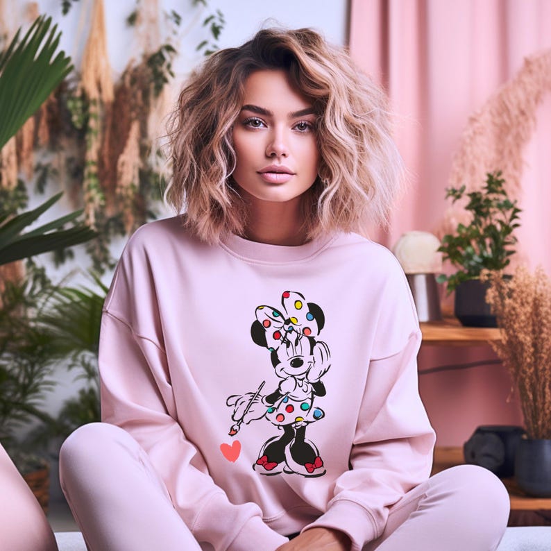May include: A pink sweatshirt with a graphic of Minnie Mouse holding a paintbrush. Minnie Mouse is wearing a red and white polka dot dress and a yellow bow with colorful polka dots.