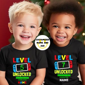 May include: Two children wearing black t-shirts with colorful video game controller designs. The shirts say "LEVEL 5 UNLOCKED" and "LEVEL 7 UNLOCKED" with space for a custom name. A circular graphic says "CUSTOMIZED NAME AND AGE."
