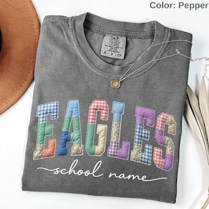 May include: A pepper-colored t-shirt featuring the word EAGLES in a multi-colored, quilted-style patchwork pattern with various floral and gingham prints. Below the word, the text school name is printed in a white, cursive script font.