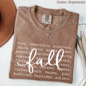May include: An espresso-colored short-sleeve t-shirt with a Comfort Colors label. The shirt features white text listing fall-themed words including cozy sweaters, scarves, jeans, boots, apple cider, pumpkin patches, crunchy leaves, giving thanks, football, harvest, candy corn, caramel apples, turkey, corn mazes, pie, bonfires, hayrides, and autumn, with the word fall written in a large white script font across the center.