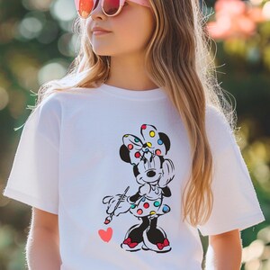 May include: A white t-shirt with a colorful graphic of Minnie Mouse painting a heart. Minnie Mouse is wearing a red and white polka dot dress and a colorful polka dot bow.