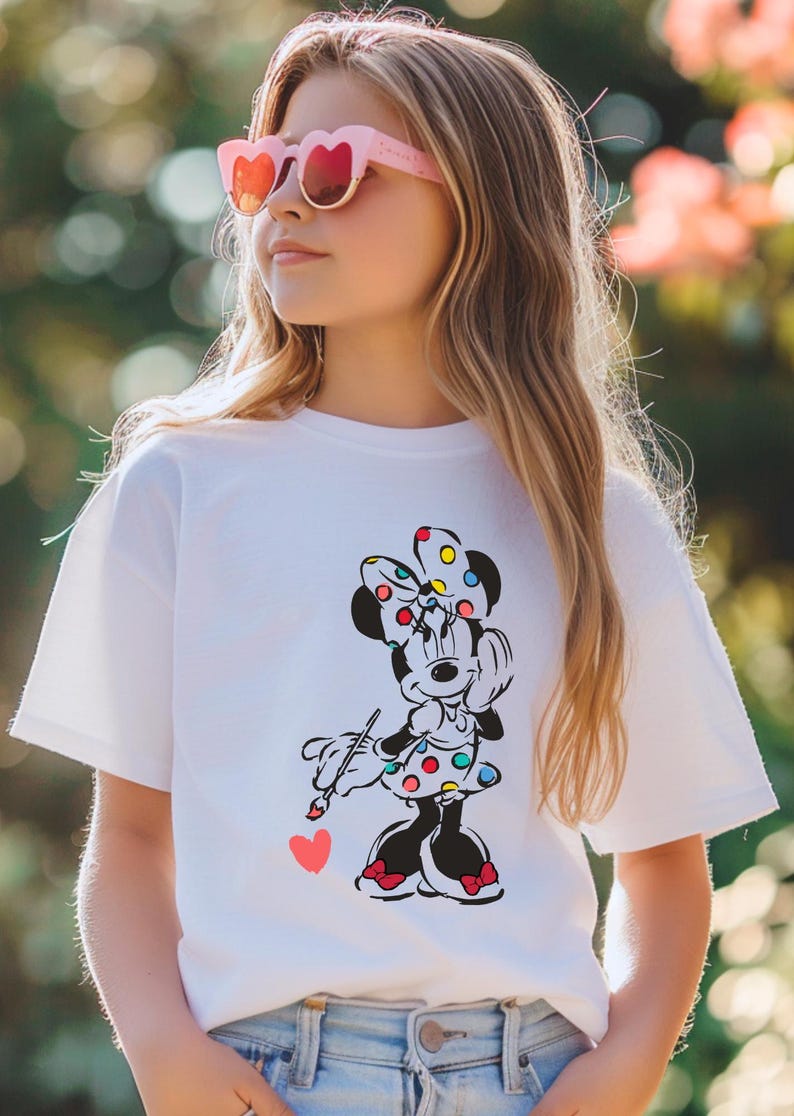May include: A white t-shirt with a colorful graphic of Minnie Mouse painting a heart. Minnie Mouse is wearing a red and white polka dot dress and a colorful polka dot bow.