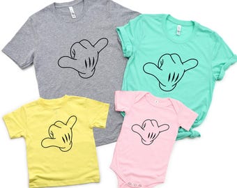May include: A set of four t-shirts featuring a black cartoon hand making the shaka sign. The shirts are in grey, turquoise, yellow, and pink.