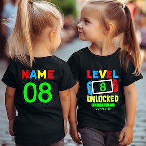 May include: Two children wearing black t-shirts with colorful text. The front shirt says "LEVEL 8 UNLOCKED" with a video game controller graphic. The back shirt says "NAME 08".