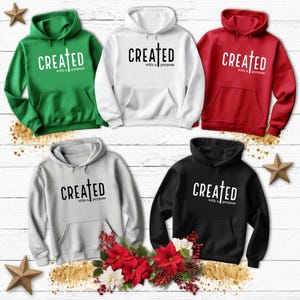 May include: Five hooded sweatshirts in different colors, each with the text "CREATED with a purpose" and a cross symbol printed on the front. The sweatshirts are displayed on a wooden background with Christmas decorations.
