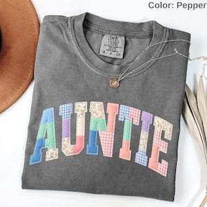 May include: Pepper-colored t-shirt with the word "AUNTIE" in colorful patchwork letters. The shirt has a crew neck and short sleeves. A silver necklace with a pendant is draped across the shirt.
