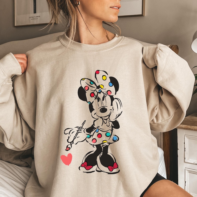 May include: A beige sweatshirt with a graphic print of Minnie Mouse holding a paintbrush and a heart. Minnie Mouse is wearing a red bow with colorful polka dots and a polka dot dress.