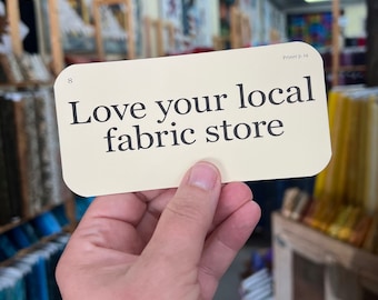 May include: A hand holds a cream-colored card with the text "Love your local fabric store" in black. The card is in a fabric store with shelves of colorful fabrics in the background.