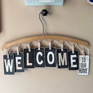 May include: A wooden hanger with the word "WELCOME" spelled out in black letters on individual cards hanging from it. A smaller card with the text "TO OUR HOME" hangs below the word "WELCOME".