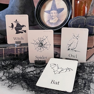 May include: Halloween-themed cards featuring a witch on a broom, a spider in a web, an owl, and a bat. The cards are cream-colored with black illustrations and text. A vintage portrait of a person wearing a witch hat is in the background.