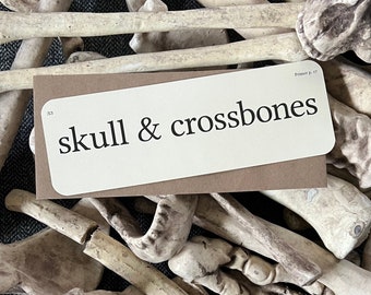 May include: A white card with brown backing, featuring the text "skull & crossbones" in black font. The card is surrounded by a pile of white plastic bones.