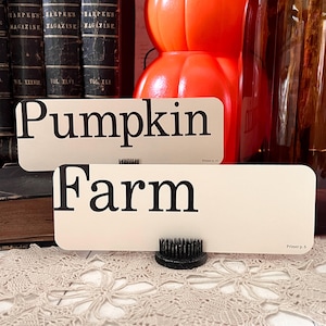 May include: Two cream-colored rectangular signs with the words "Pumpkin" and "Farm" in bold black letters. The signs are propped up by a black floral frog. The background includes vintage books, orange pumpkins, and glass bottles.