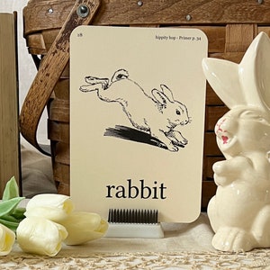 May include: A black and white illustration of a rabbit hopping on a white card with the word "rabbit" printed in black. The card is labeled "hippity hop - Primer p. 34" and has the number 18 in the top right corner.